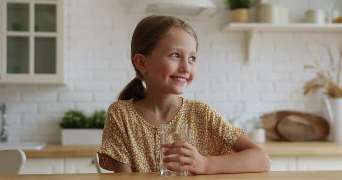 Thirsty little girl sit at table in kitchen holding glass drinking fresh cool filtered clear aqua, preventing dehydration. Healthy routine, water purification system at home, body refreshment concept