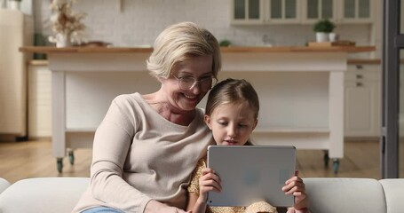 Older 50s granny and little granddaughter resting on couch holding tablet modern wireless device, web surfing internet, making purchase online, having fun with electronic gadget and e services concept - Powered by Adobe