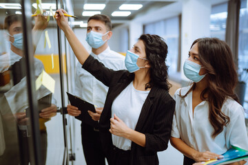 Business colleagues  in protective face mask writing on sticky note on glass wall with coworkers standing by in office. Startup Business. Planning, analysis. Covid-2019.