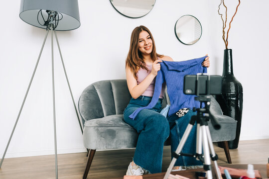 Attractive caucasian young woman video blogger showing new clothing via her blog on social media, holding blouse and looking at  smartphone camera fixed on tripod.