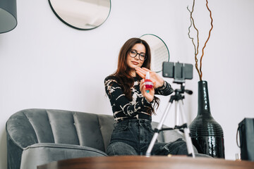 Attractive caucasian young woman video blogger showing beauty products via her blog on social media, looking at  smartphone camera fixed on tripod.