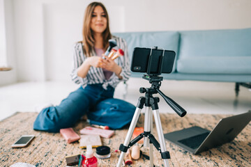Attractive caucasian young woman video blogger showing beauty products via her blog on social media, holding makeup brushes looking at  smartphone camera fixed on tripod.
