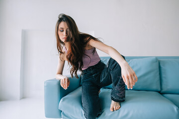 Obraz premium Attractive stylish caucasian woman with long brunette hair posing on blue sofa in modern living room with white walls.