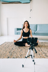 Beautiful brunette fitness woman meditate in front camera on tripod, doing yoga at home. Staying fit and healthy