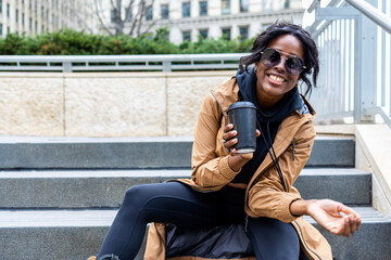 African American black woman is in downtown Chicago enjoying the weather drinking hot coffee. while touring the city architecture. She wears a fashionable brown trench coat with a happy smile