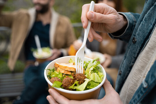 Young people eating together takeaway food outdoors in the park. Closeup on the chicken salad into plastic free bowls and compostable cutlery. New normal habits concept.