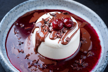 Modern style traditional blancmange almond pudding with cherry, chocolate cream and sour cherry gelee compote coulis served as close-up in a Nordic design plate on a black board