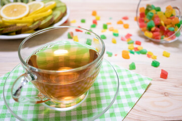 Glass cup of tea, multicolored candied fruits, a plate with lemon and kiwi slices on a wooden background with white and green napkins close-up