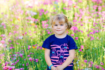 Portriat of adorable, charming toddler girl in flowers meadow. Smiling happy baby child on summer day with colorful flowers, outdoors. Happiness and summertime.