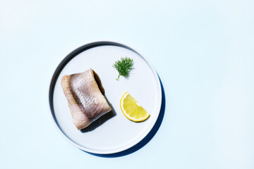 Herring fillet rolled up with lemon slice and dill on the plate on light blue background. Minimal...