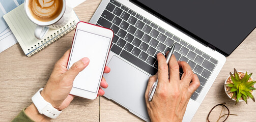 Man's hand using smartphone on wooden table with laptop, book and coffee