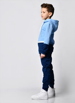 Side View Kid In Blue Warm Overalls, White Sneakers Walking Along The Studio Wall. Full-length Portrait. Children's Fashion, Casual Wear, Comfortable Children Clothing For Winter And Autumn