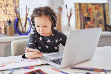 Cute small preschool girl in headphones sitting at a table using a laptop computer and smartphone for education or homework doing. Doing business or small businesswoman concept. High quality photo © boytsov