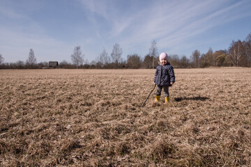 Naklejka na ściany i meble a little girl runs and plays in the field in the spring. a child in rubber boots plays with a stick