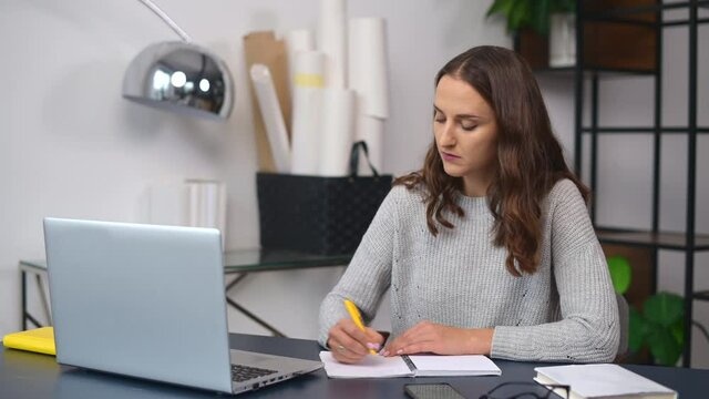 Concentrated business woman is using laptop computer for watching online webinars, female employee sits on the workplace in the office and writing down notes with a pen, woman studying online