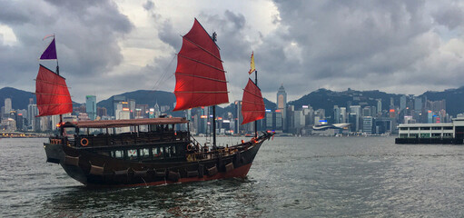Traditional Chinese junk with red sails in Victoria Harbor, Hong Kong in China with panoramic city...