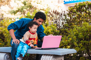 black young father with his son, on a video call with a pink computer, outdoors, father's day concept