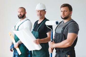 Crew of three professional builder with painting roll and renovation plans standing in empty...