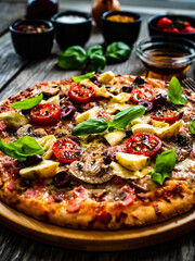 Pizza capricciosa with white mushrooms, ham, artichoke, tomatoes, olives, parmesan and mozzarella on wooden background
