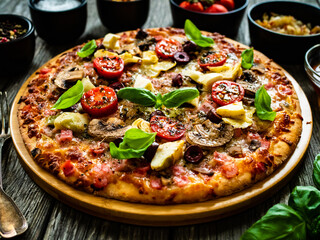 Pizza capricciosa with white mushrooms, ham, artichoke, tomatoes, olives, parmesan and mozzarella on wooden background
