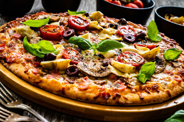 Pizza capricciosa with white mushrooms, ham, artichoke, tomatoes, olives, parmesan and mozzarella on wooden background
