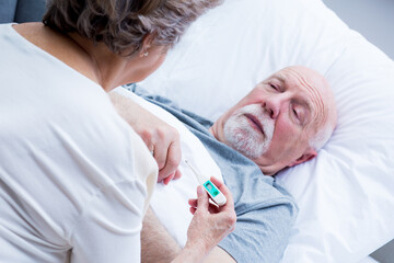 Elderly wife measures the temperature of her sick husband lying in bed