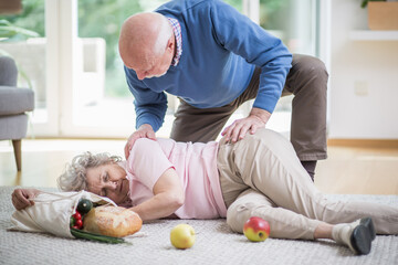 Senior woman lying on the floor of her apartment, and her husband helping her to get up