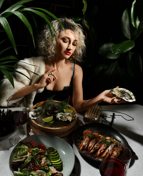 Rich wealthy woman in black dress has a dinner in seafood restaurant sits holding oysters, prawns, mussels in hand over dark background