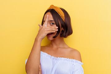 Young mixed race woman isolated on yellow background blink at the camera through fingers, embarrassed covering face.