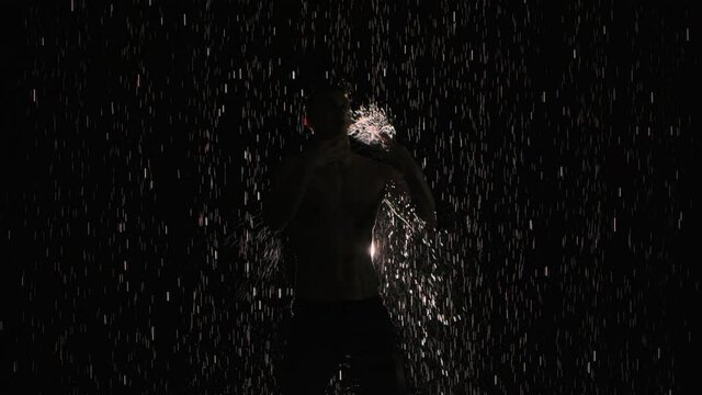 Silhouette of wet male dancer performing modern ballet dance in rain and splashing water. Male sensual modern choreography on black background in center of light beam. Slow motion. Close up.