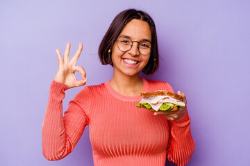 Young mixed race woman holding a sandwich isolated on purple background cheerful and confident showing ok gesture.