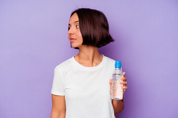 Young mixed race holding a water bottle isolated on purple background looks aside smiling, cheerful and pleasant.
