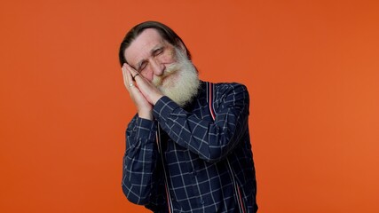 Tired elderly bearded gray-haired man going to sleep laying down on his palms, yawning, dozing off during day, taking nap and resting, comfortable bedtime. Senior old grandfather on orange background