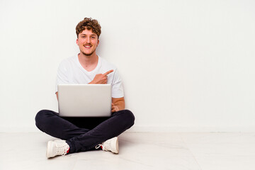 Young caucasian man sitting on the floor holding on laptop isolated on white background smiling and pointing aside, showing something at blank space.
