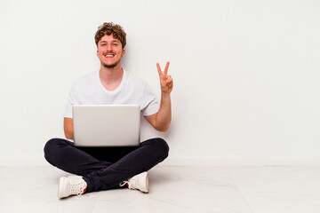 Young caucasian man sitting on the floor holding on laptop isolated on white background showing victory sign and smiling broadly.