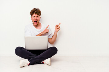 Young caucasian man sitting on the floor holding on laptop isolated on white background pointing with forefingers to a copy space, expressing excitement and desire.