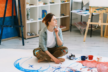 Hand painting. Art therapy. Creative hobby. Relaxing leisure. Inspired peaceful pensive female artist sitting cross-legged on colorful abstract artwork on floor in modern workshop.