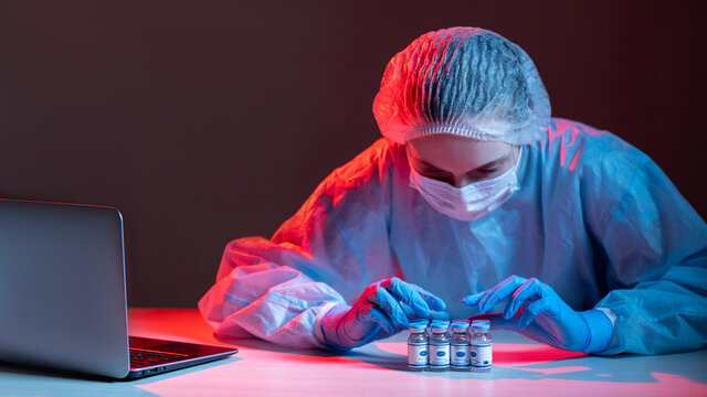 Vaccine analysis. Covid-19 immunization. Clinic research. Female doctor in ppe face mask gloves checking vial dose bottles at workplace with laptop in red blue neon light isolated on dark background.