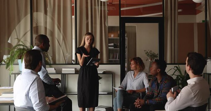 Smiling young female businesswoman holding digital computer tablet in hands, presenting research results to interested multiracial colleagues or giving educational workshop to teammates in office.