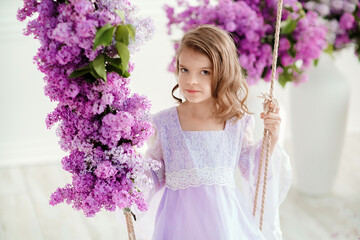 Beautiful little girl of preschool age in a delicate dress sitting on a swing decorated with flowers of lilac.
