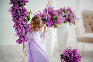 Beautiful little girl of preschool age in a delicate dress sitting on a swing decorated with flowers of lilac.