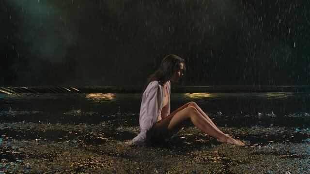 A young woman performs modern ballet choreography while sitting on the surface of the water in the rain. A flexible female body wriggles in a dark studio with beautiful light. Slow motion. Close up.