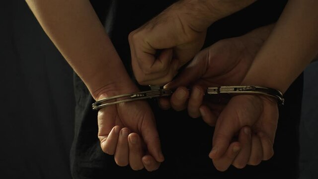 A girl in a black T-shirt stands with her back to the camera. The handcuffs are removed from her hands. On a black background