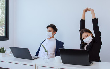 Two asian business man and woman wearing formal suit, face mask to protect virus, stretching and taking break while working in indoor office or workplace with labtops on white table.
