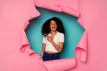 Young African American woman in torn paper isolated on blue background smiling and pointing aside, showing something at blank space.