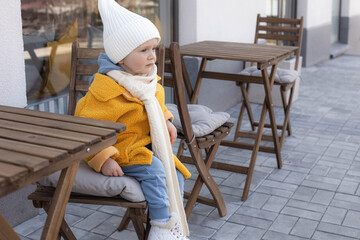 Small smiling baby girl in yellow jacket near the table in outdoor cafe