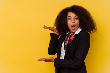 Young African American air hostess isolated on yellow background shocked and amazed holding a copy space between hands.