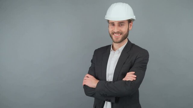 Three-quarters portrait of a smiling business man in a business suit and white hard hat isolated on gray background folding hands on chest 4K