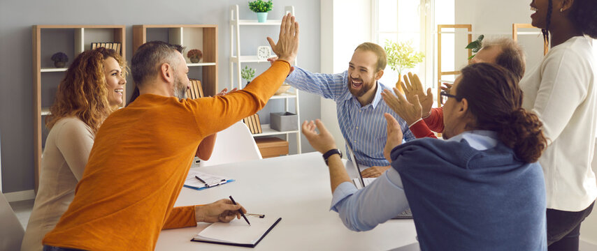 Happy successful multiracial business team clapping hands while two colleagues giving high fives gesture each other. Businesspeople laugh and cheer sitting at table in office on briefing meeting