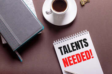 On a brown background, diaries, a white cup of coffee and a notebook with the text TALENTS NEEDED.